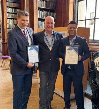 Three men in business attire holding certificates in a library setting with bookshelves in the background