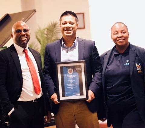 Three people in business attire posing together indoors, with the center man holding a framed award or certificate